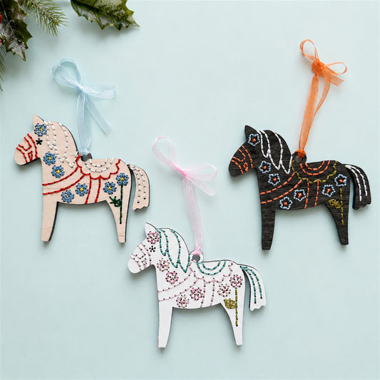 Three Dala horse embroidery ornaments in light, white and dark wood with ribbons on a pale blue background
