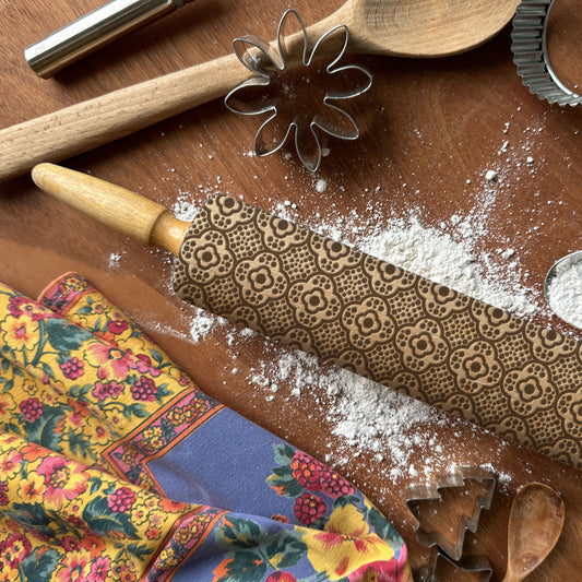 Patterned rolling pin with flour, wooden spoon, and floral towel on a wooden surface.