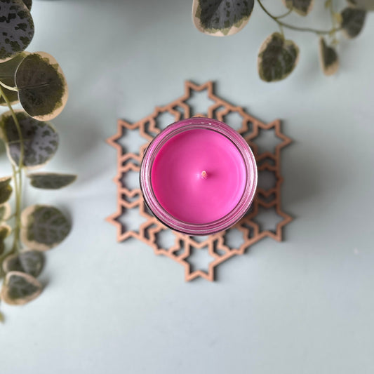 Pink candle on a decorative trivet on a white surface with green leaves in the background