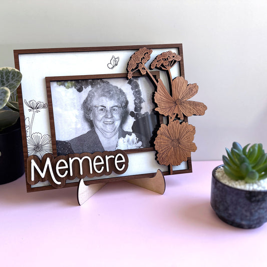 Decorative photo frame with a black and white photo of an elderly woman, wooden butterflies, and the word 'Memere' on a light pink surface.