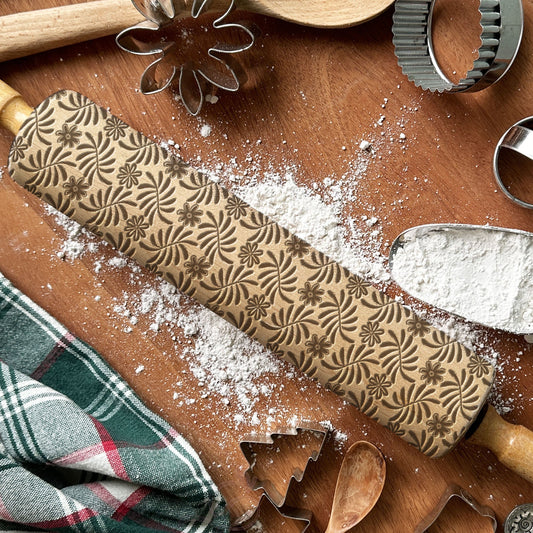 Wooden rolling pin with embossed patterns on a wooden surface with flour and cookie cutters.