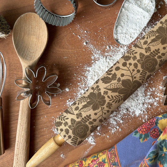 Baking tools including a wooden spoon, rolling pin with floral patterns, and cookie cutters on a wooden surface with flour.