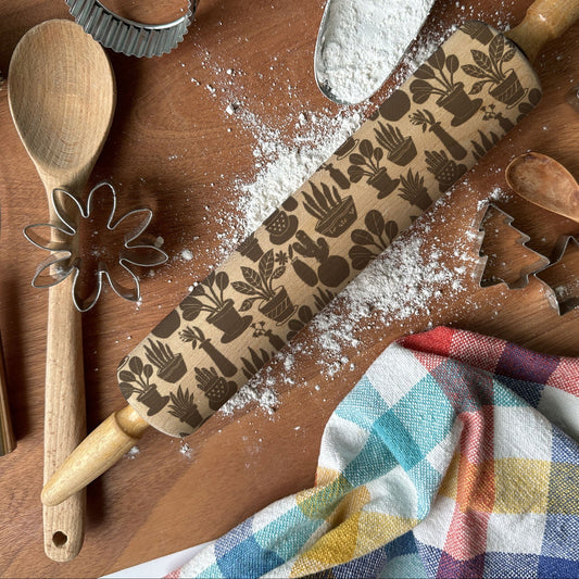 Baking tools including a rolling pin with embossed designs, wooden spoon, whisk, and cookie cutters on a wooden surface.