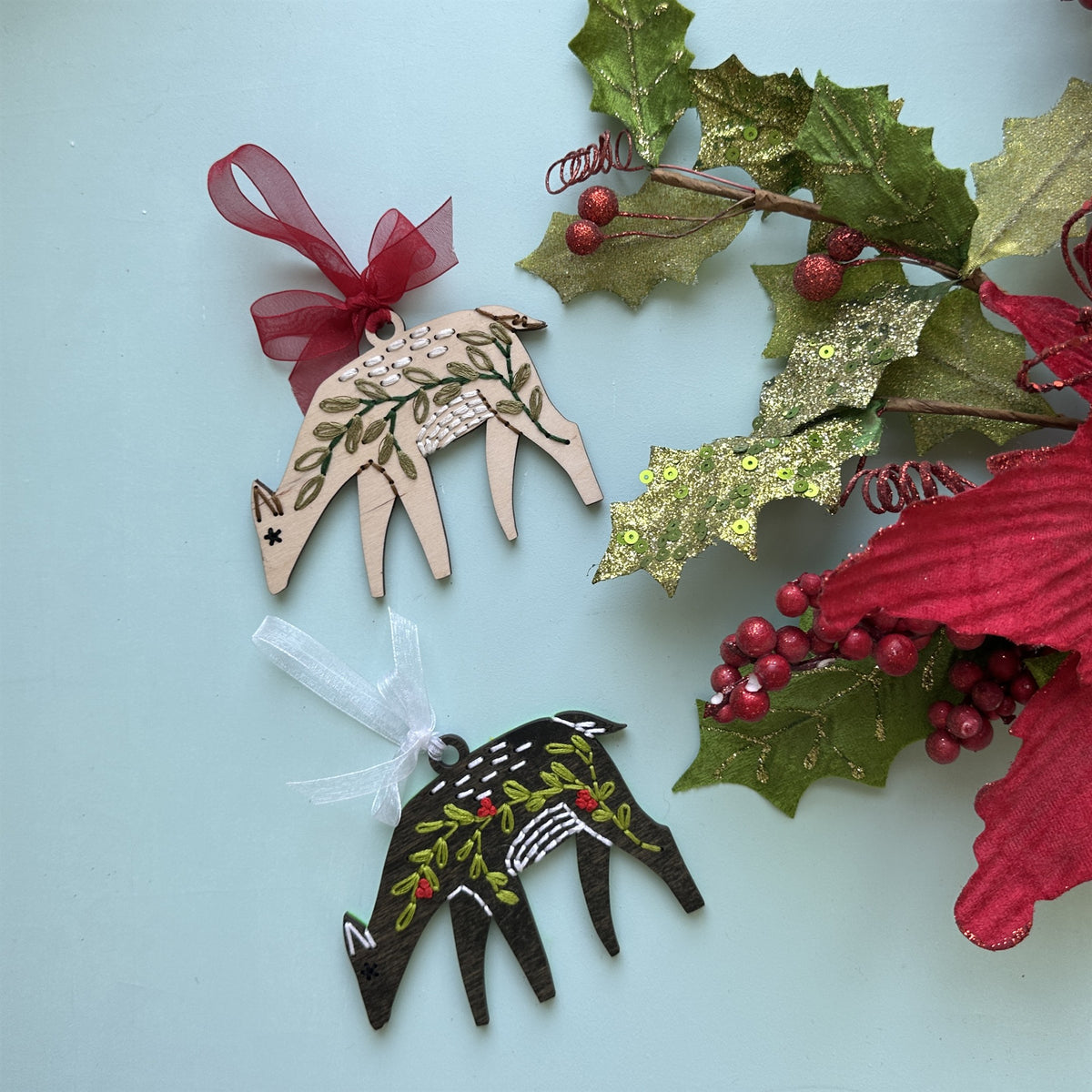 Light wood and dark stained deer embroidery ornaments with holly and a red poinsettia on a pale blue background