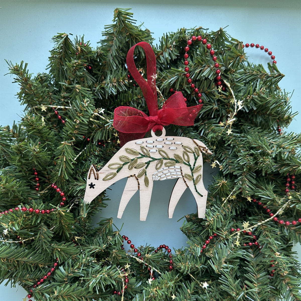 Light wood deer embroidery ornament with a red bow hanging in an evergreen Christmas wreath