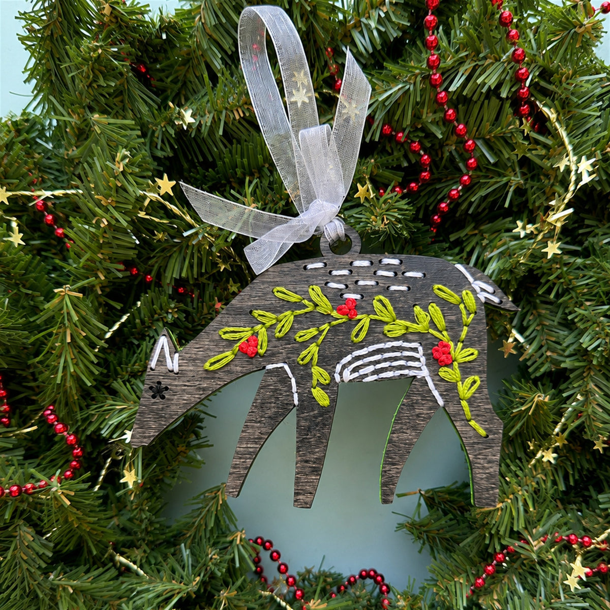 Gray stained deer embroidery ornament with green and red stitching and a white ribbon in an evergreen wreath