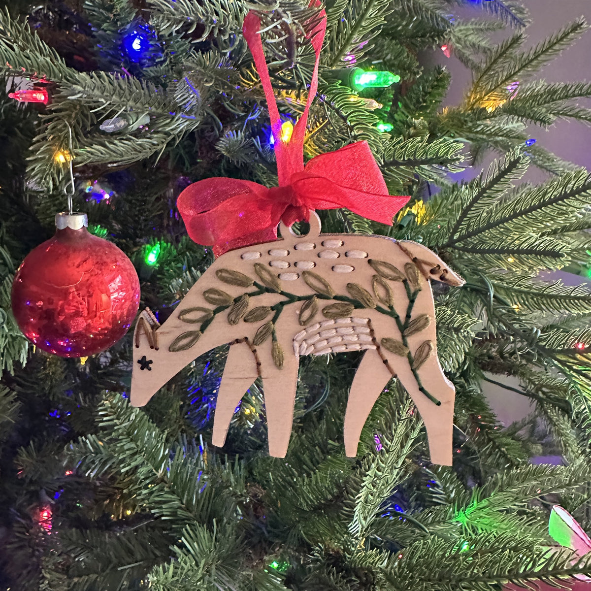 Light wood deer embroidery ornament with a red bow hanging on a lit Christmas tree