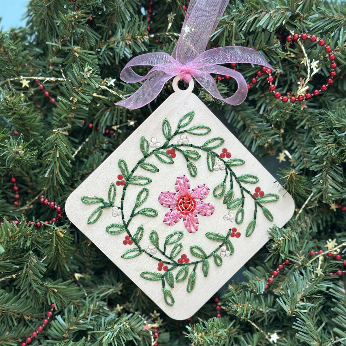Light wood floral wreath embroidery ornament with a pink ribbon hanging in an evergreen Christmas wreath