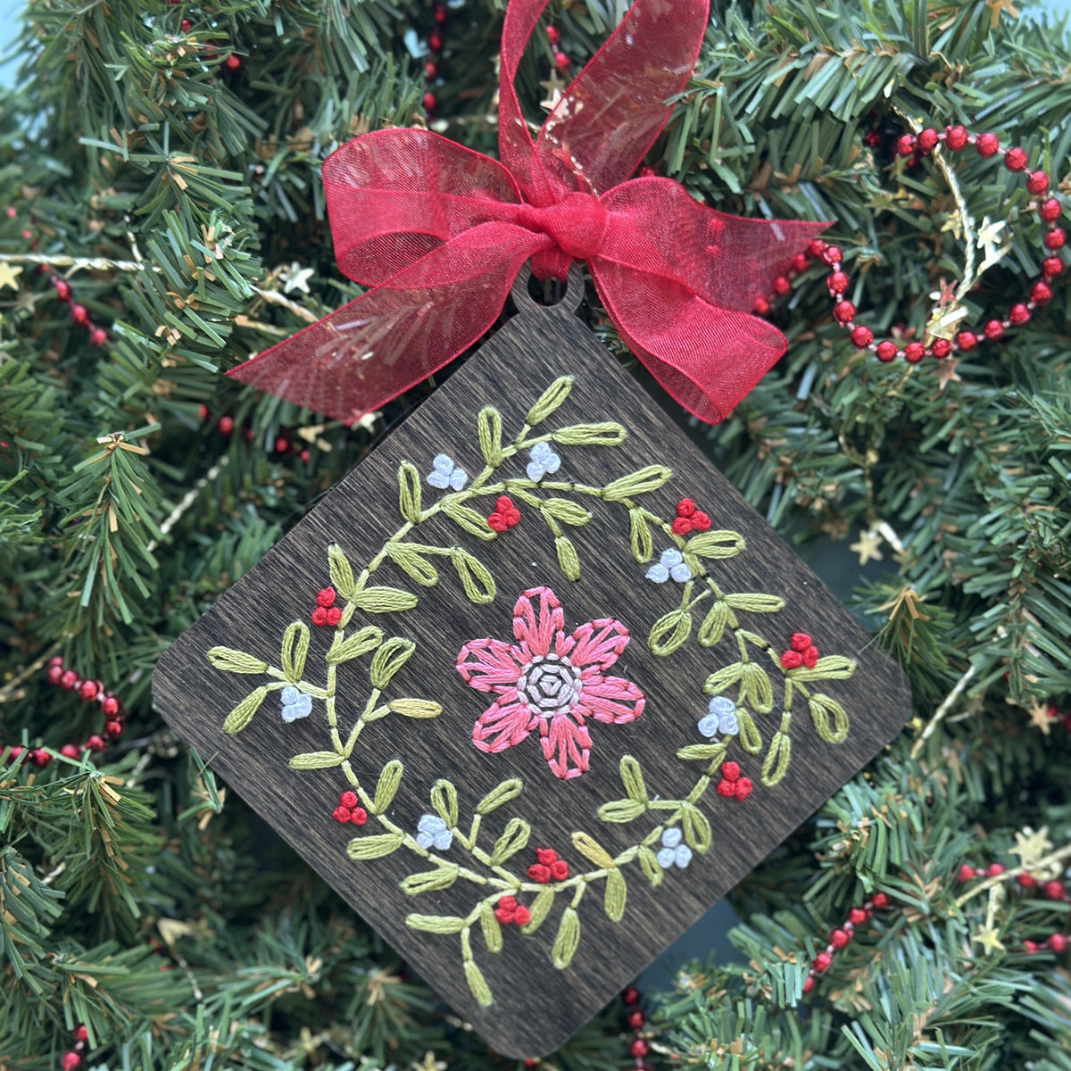 Dark stained floral wreath embroidery ornament with a red ribbon hanging in an evergreen wreath