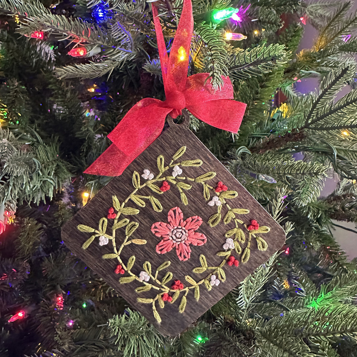 Dark stained floral wreath embroidery ornament with a red ribbon on a lit Christmas tree
