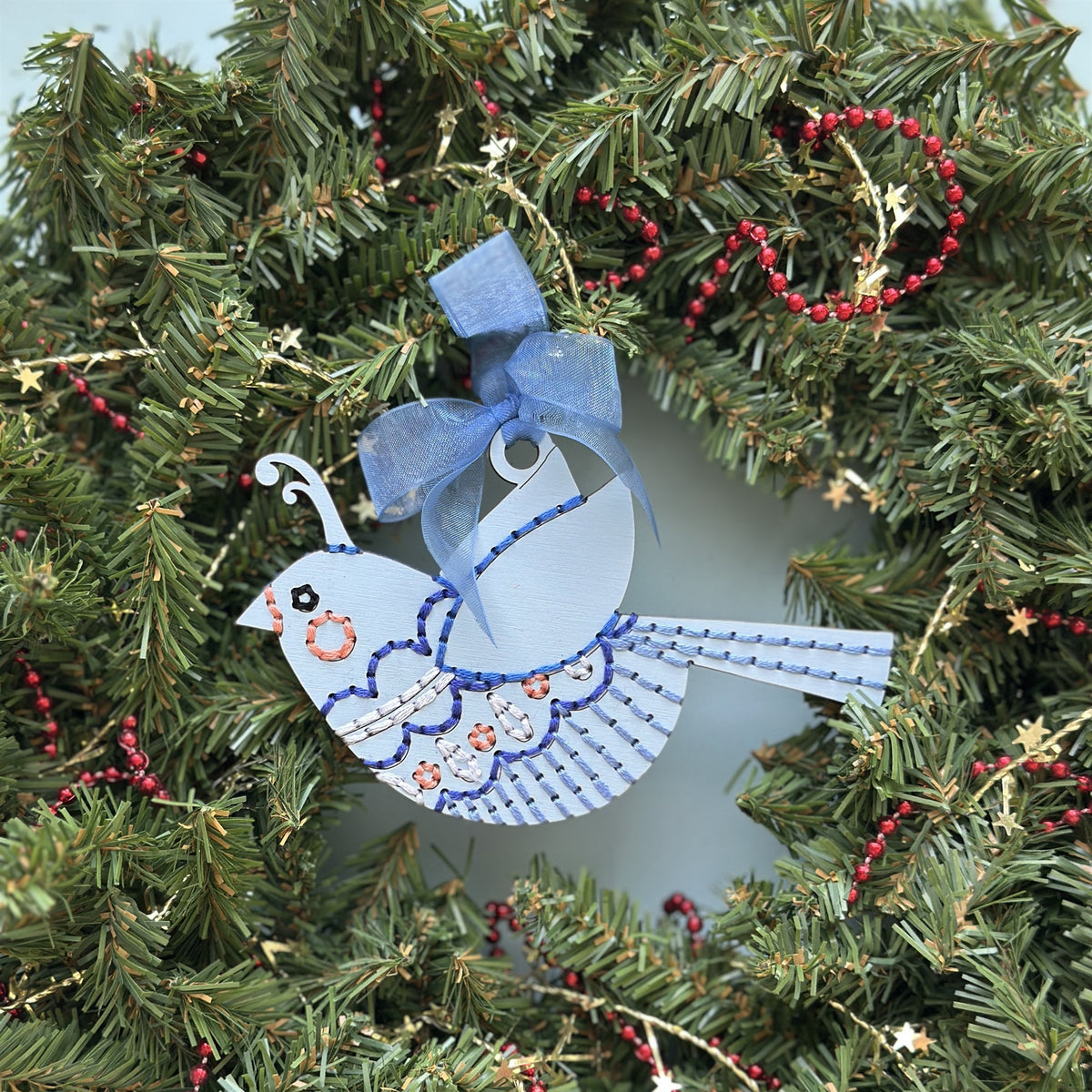Pale blue songbird embroidery ornament with a blue ribbon hanging in an evergreen Christmas wreath