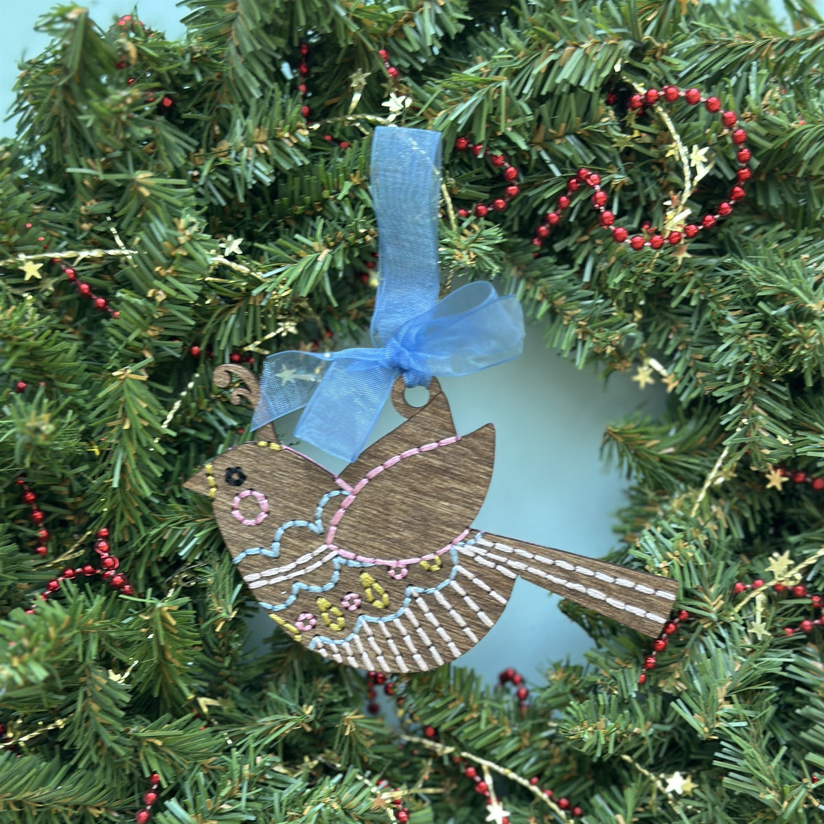 Brown stained songbird embroidery ornament with white stitching hanging in an evergreen wreath