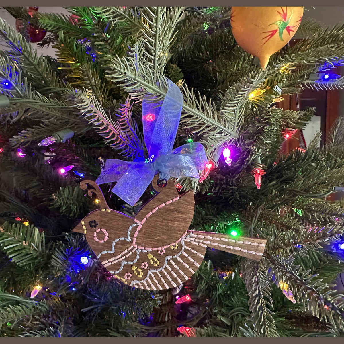 Brown stained songbird embroidery ornament with a blue bow on a lit Christmas tree