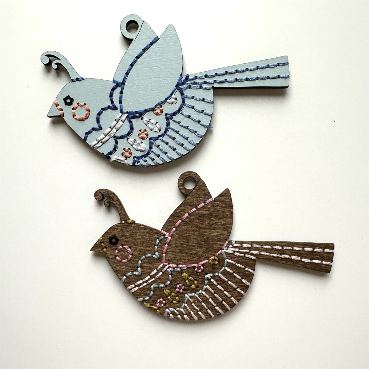 Pale blue and brown stained songbird embroidery ornaments laid flat on a white background