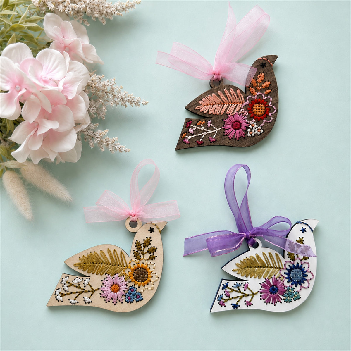 Three folk floral bird embroidery ornaments in light, white and dark wood with pink and purple ribbons