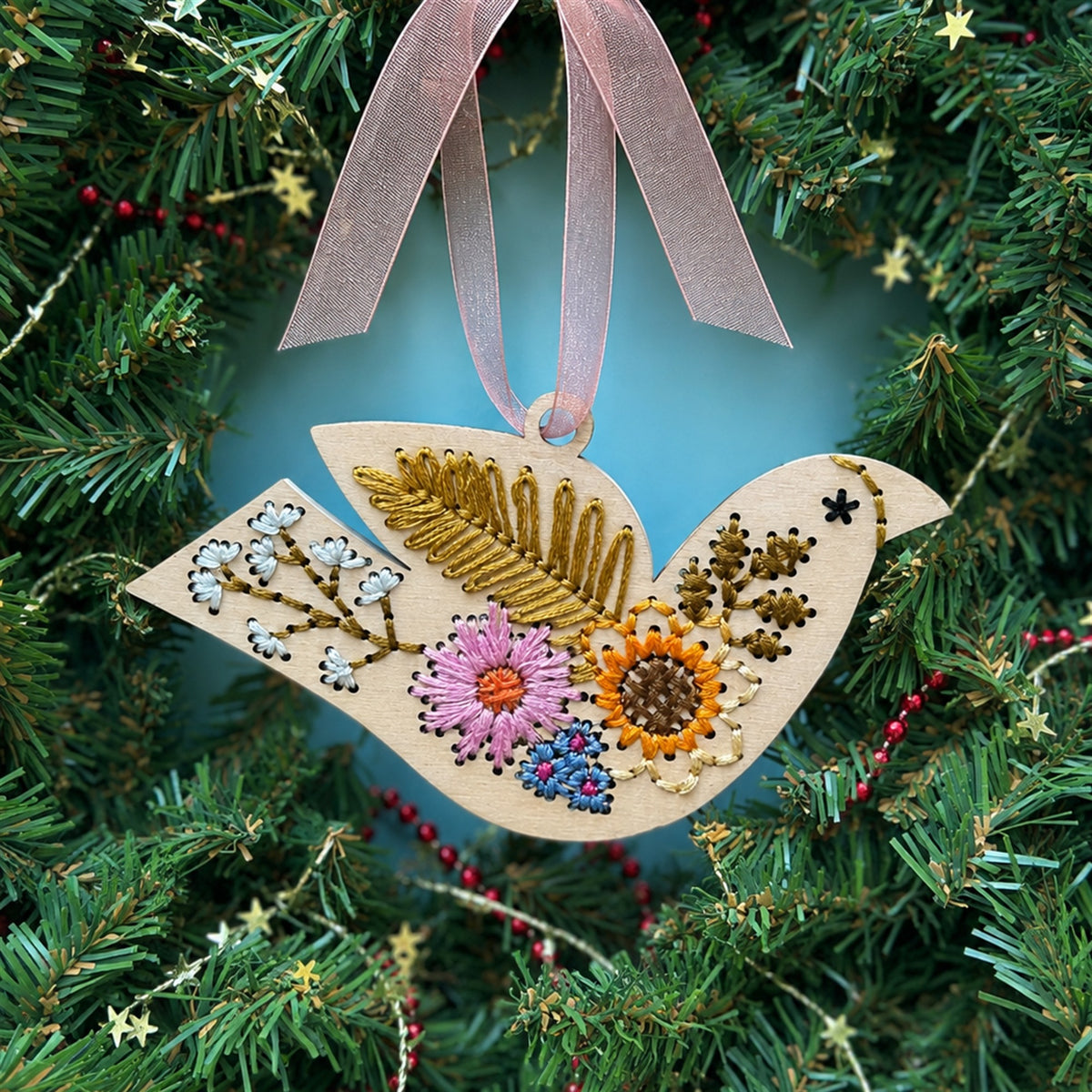 Light wood folk floral bird embroidery ornament with a pink ribbon hanging in an evergreen Christmas wreath