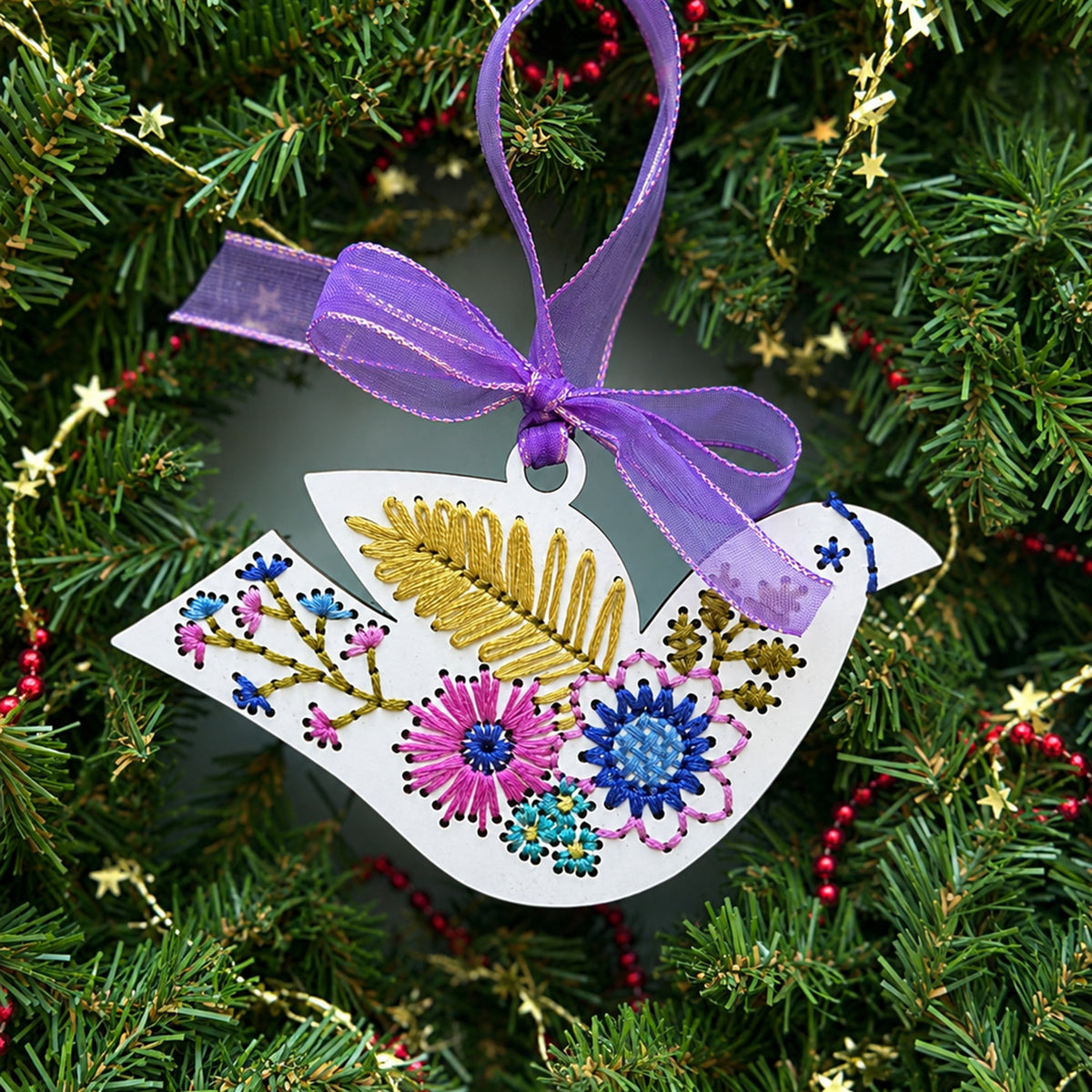 White folk floral bird embroidery ornament with a purple ribbon hanging in an evergreen wreath