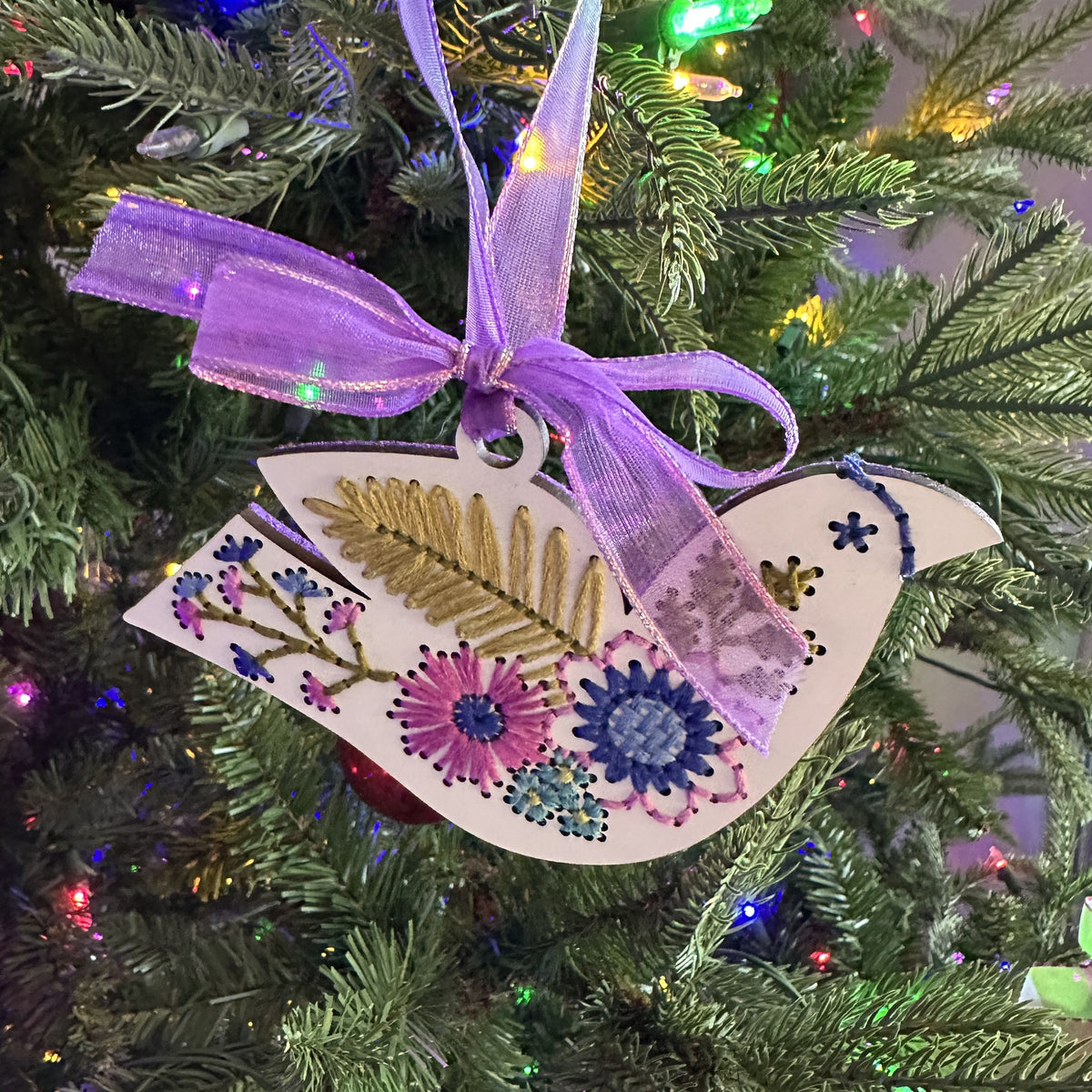 White folk floral bird embroidery ornament with a purple bow on a lit Christmas tree