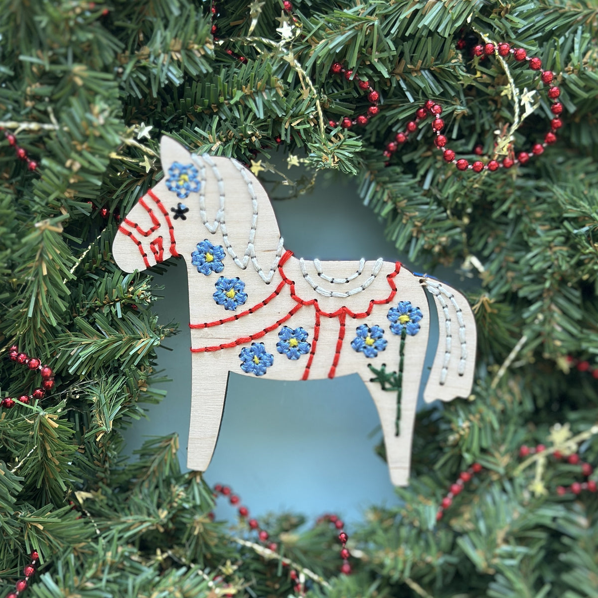 White Dala horse embroidery ornament with red and blue stitching hanging in an evergreen Christmas wreath