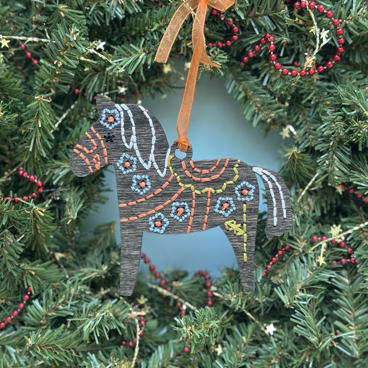 Dark stained Dala horse embroidery ornament with an orange ribbon hanging in an evergreen wreath