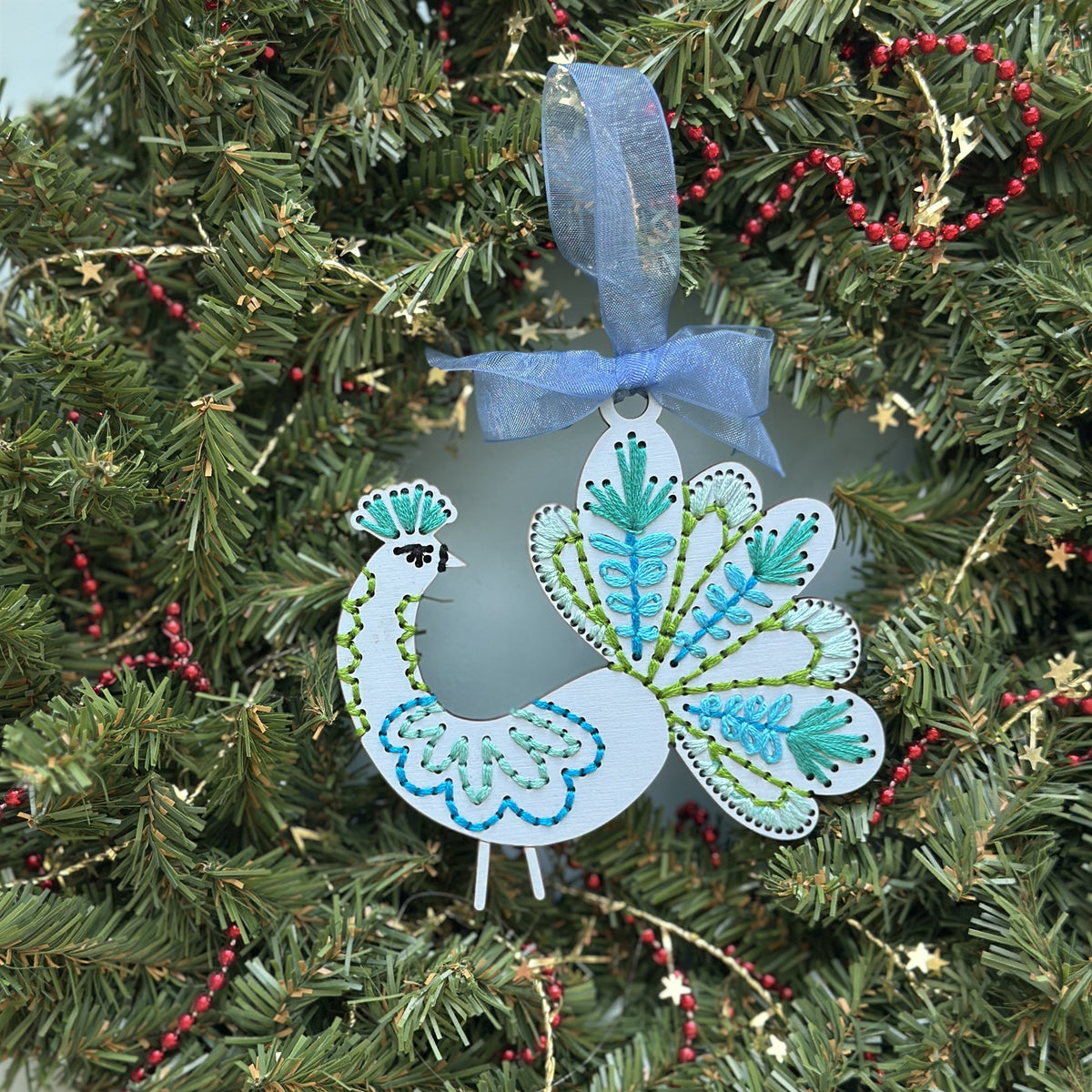 Pale blue peacock embroidery ornament with a blue ribbon hanging in an evergreen Christmas wreath