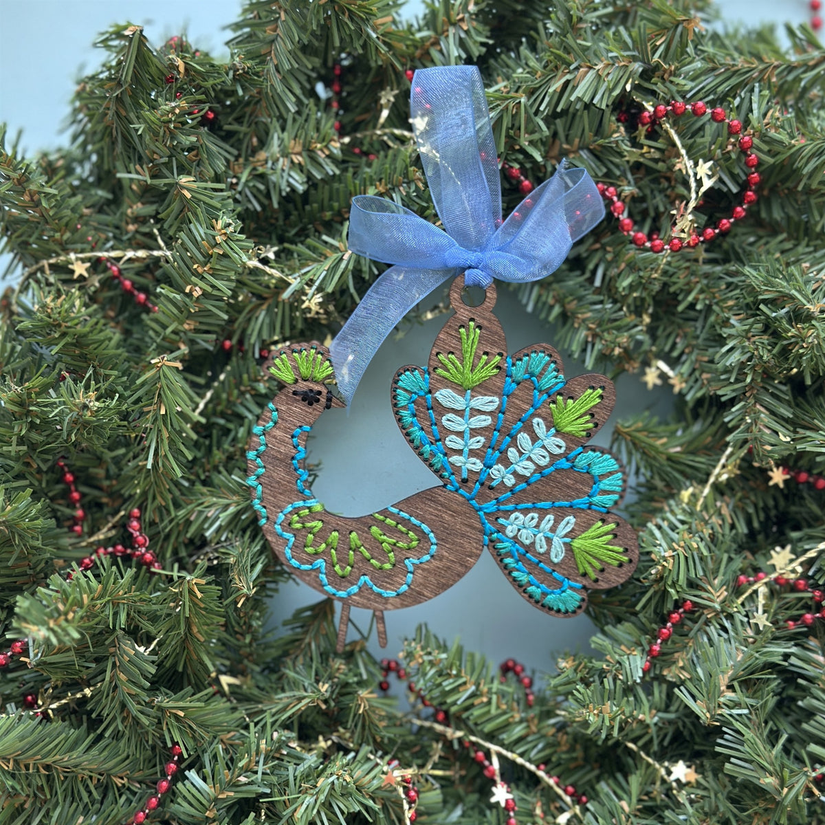 Brown stained peacock embroidery ornament with a blue ribbon hanging in an evergreen wreath