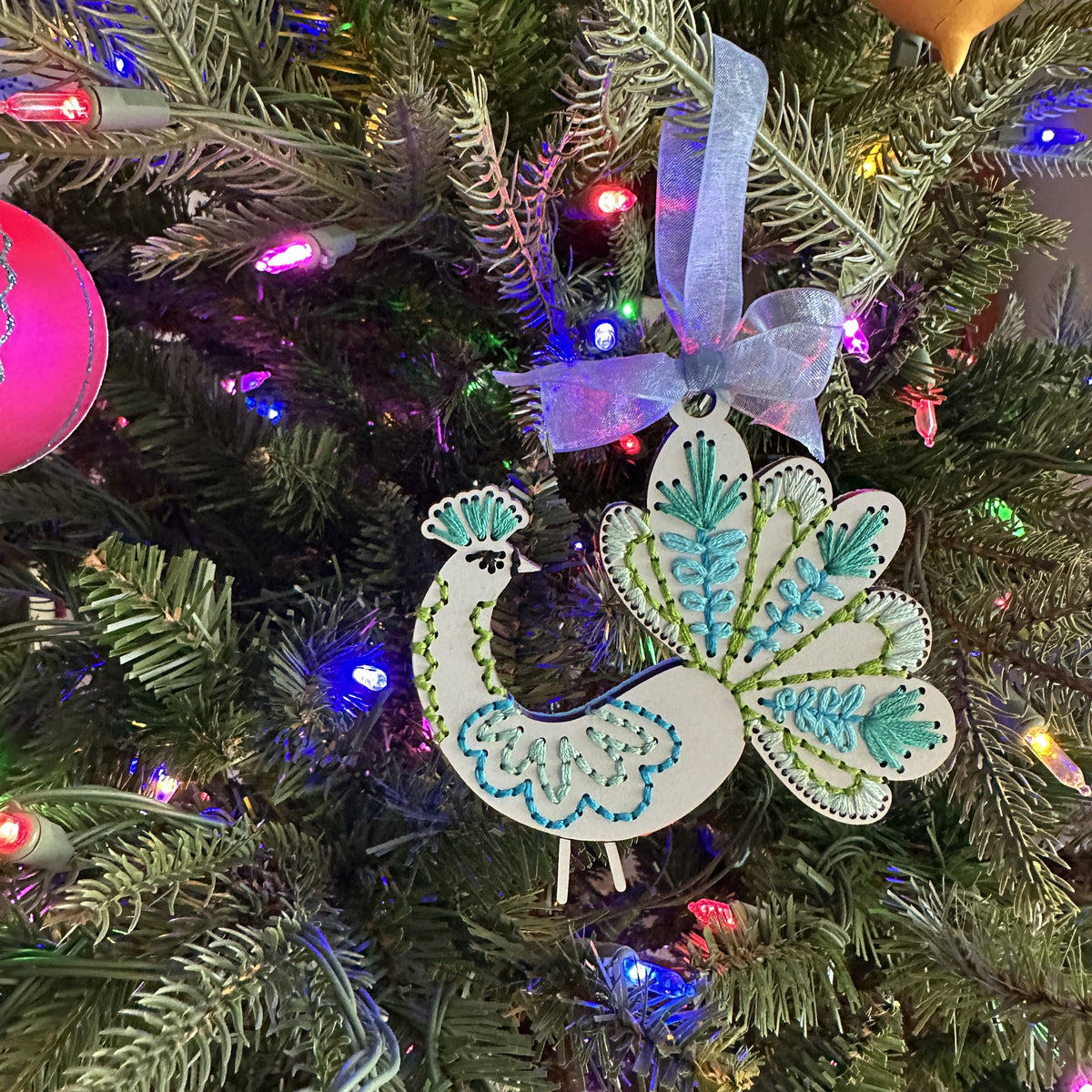 Pale blue peacock embroidery ornament hanging on a lit Christmas tree