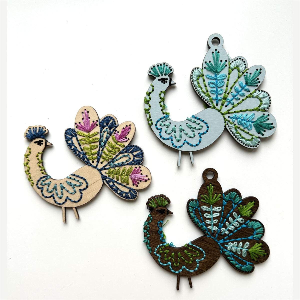 Three peacock embroidery ornaments in light, blue and dark finishes laid flat on a white background