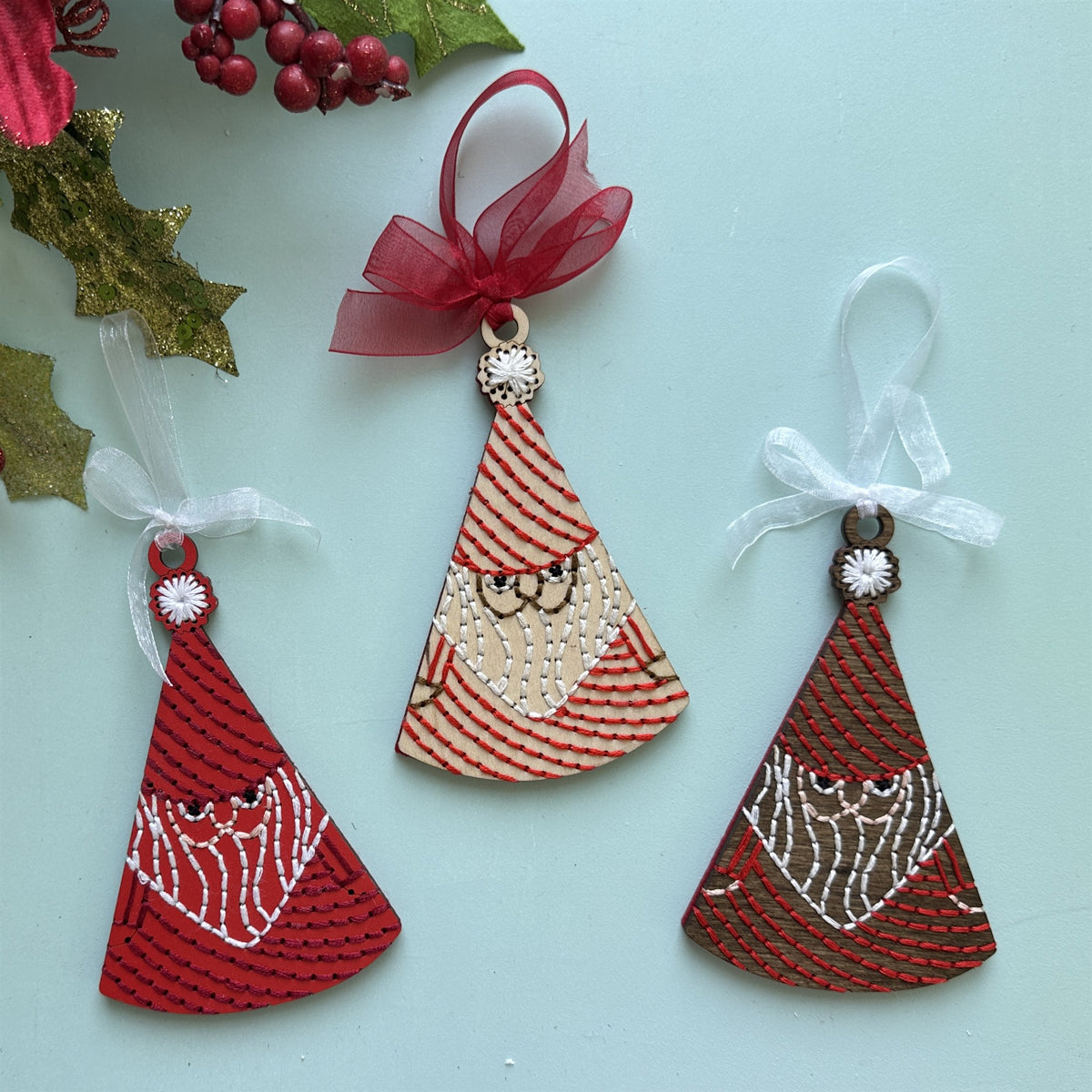Red, light wood and dark stained Santa embroidery ornaments with holly on a pale blue background
