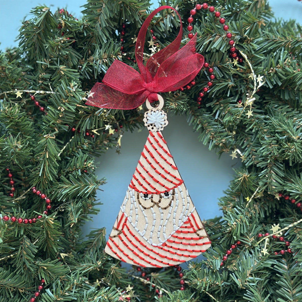 Light wood Santa embroidery ornament with a red bow hanging in an evergreen Christmas wreath