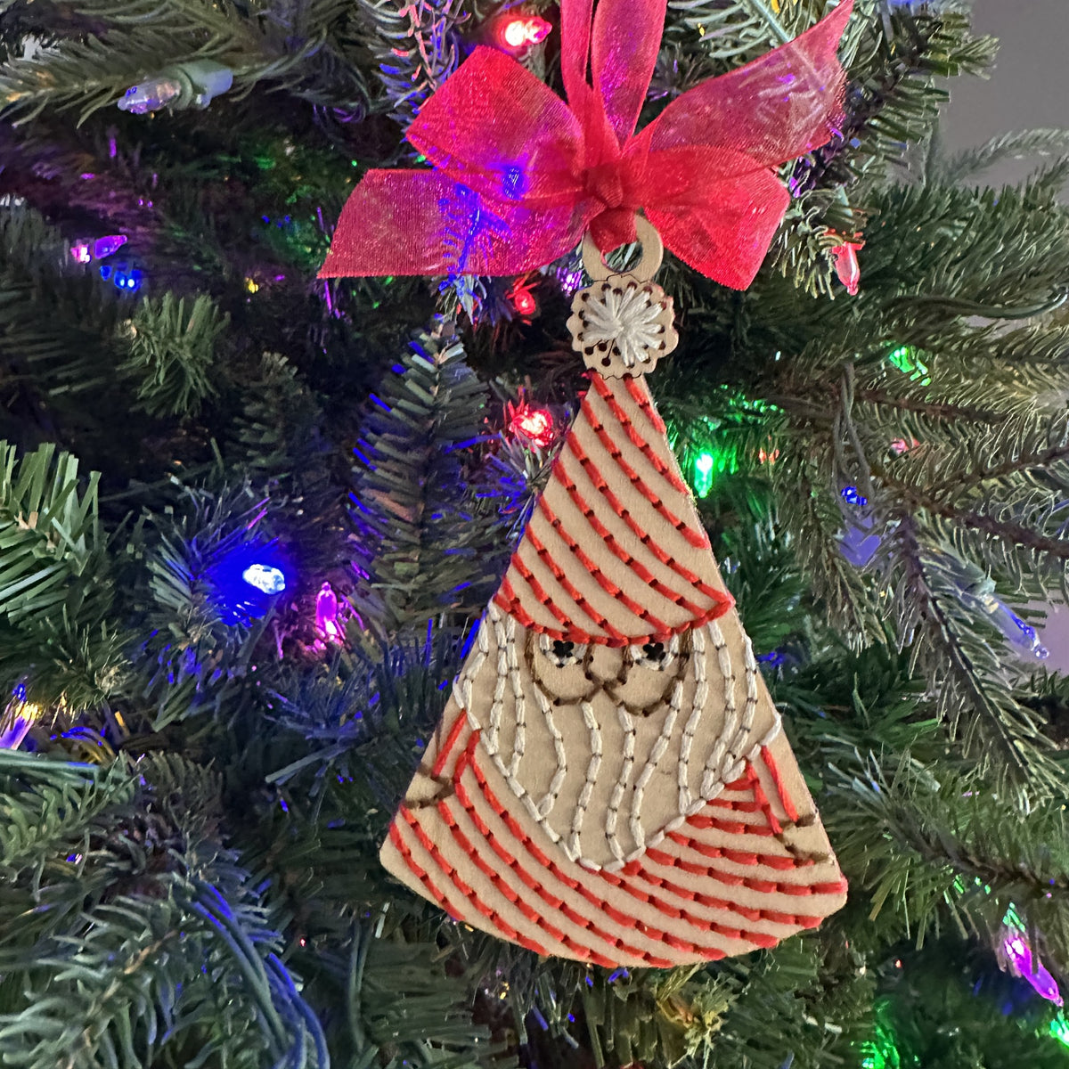 Light wood Santa embroidery ornament with a red bow on a lit Christmas tree