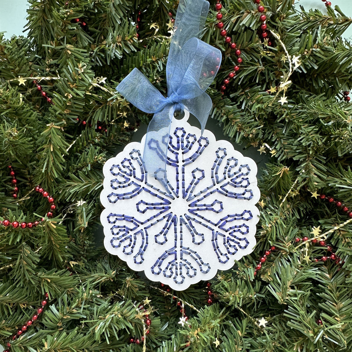 White snowflake embroidery ornament with blue stitching hanging in an evergreen Christmas wreath