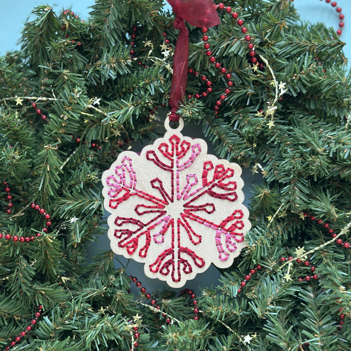 Light wood snowflake embroidery ornament with red stitching hanging in an evergreen wreath