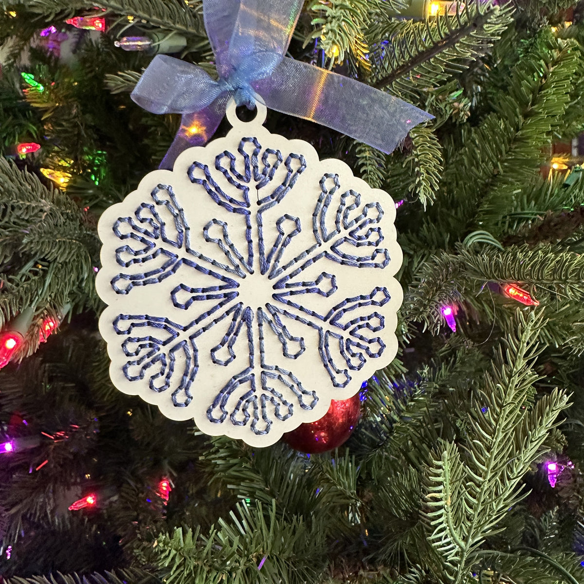 White snowflake embroidery ornament with blue stitching on a lit Christmas tree