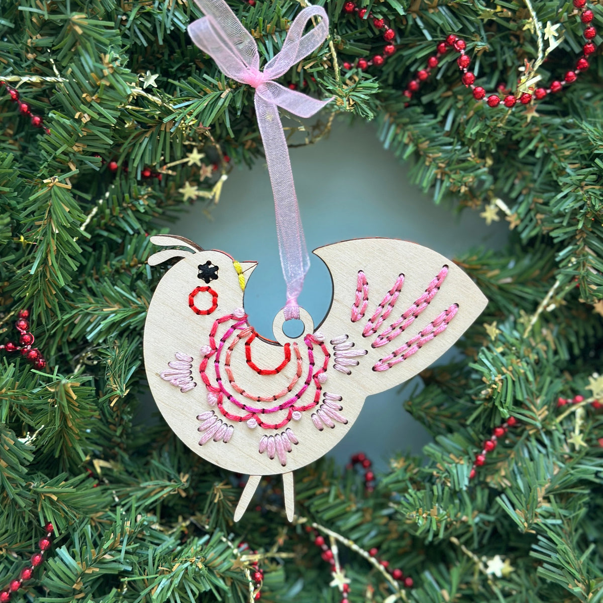 Light wood scalloped folk bird embroidery ornament with pink stitching hanging in an evergreen Christmas wreath