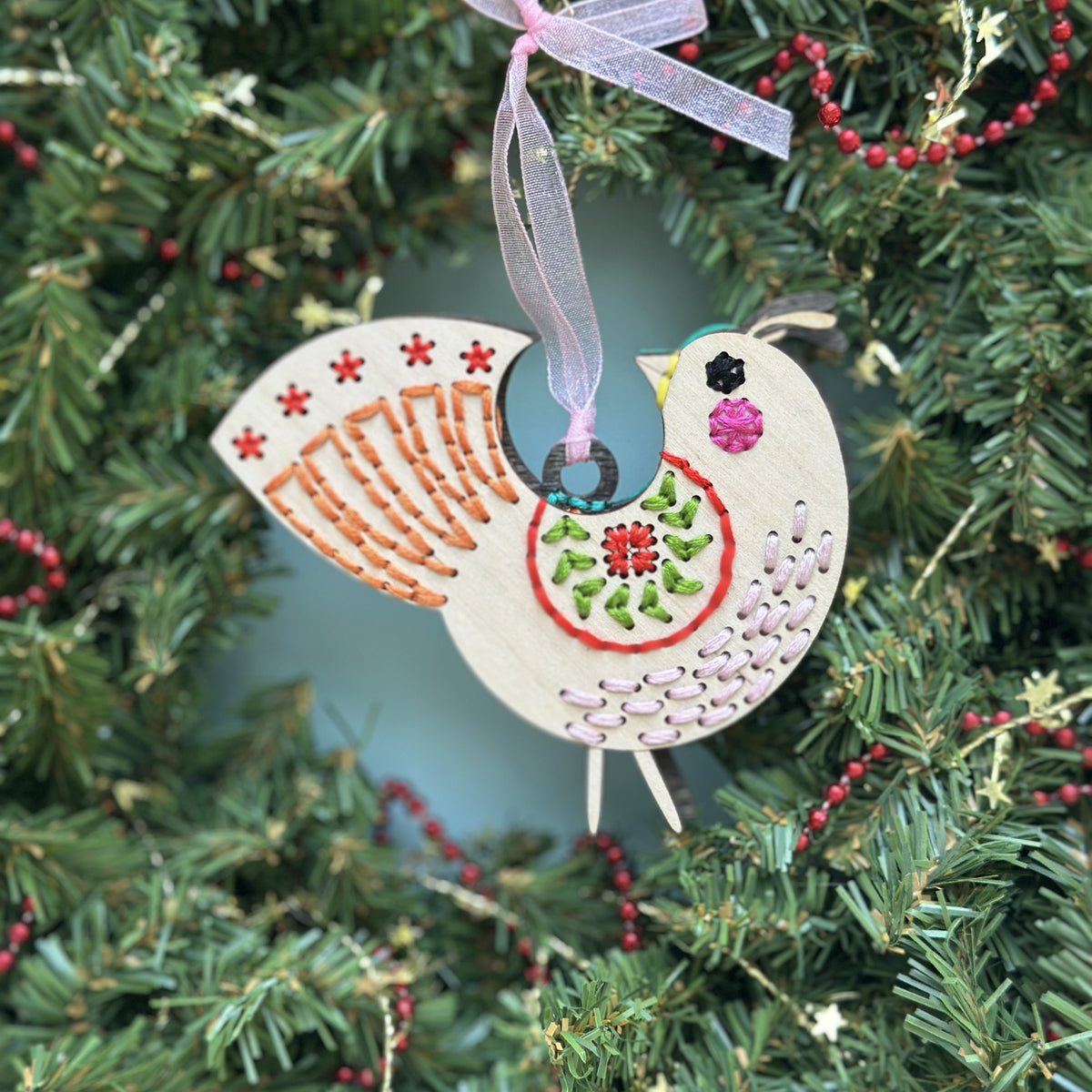 Light wood medallion folk bird embroidery ornament with a pink ribbon hanging in an evergreen Christmas wreath