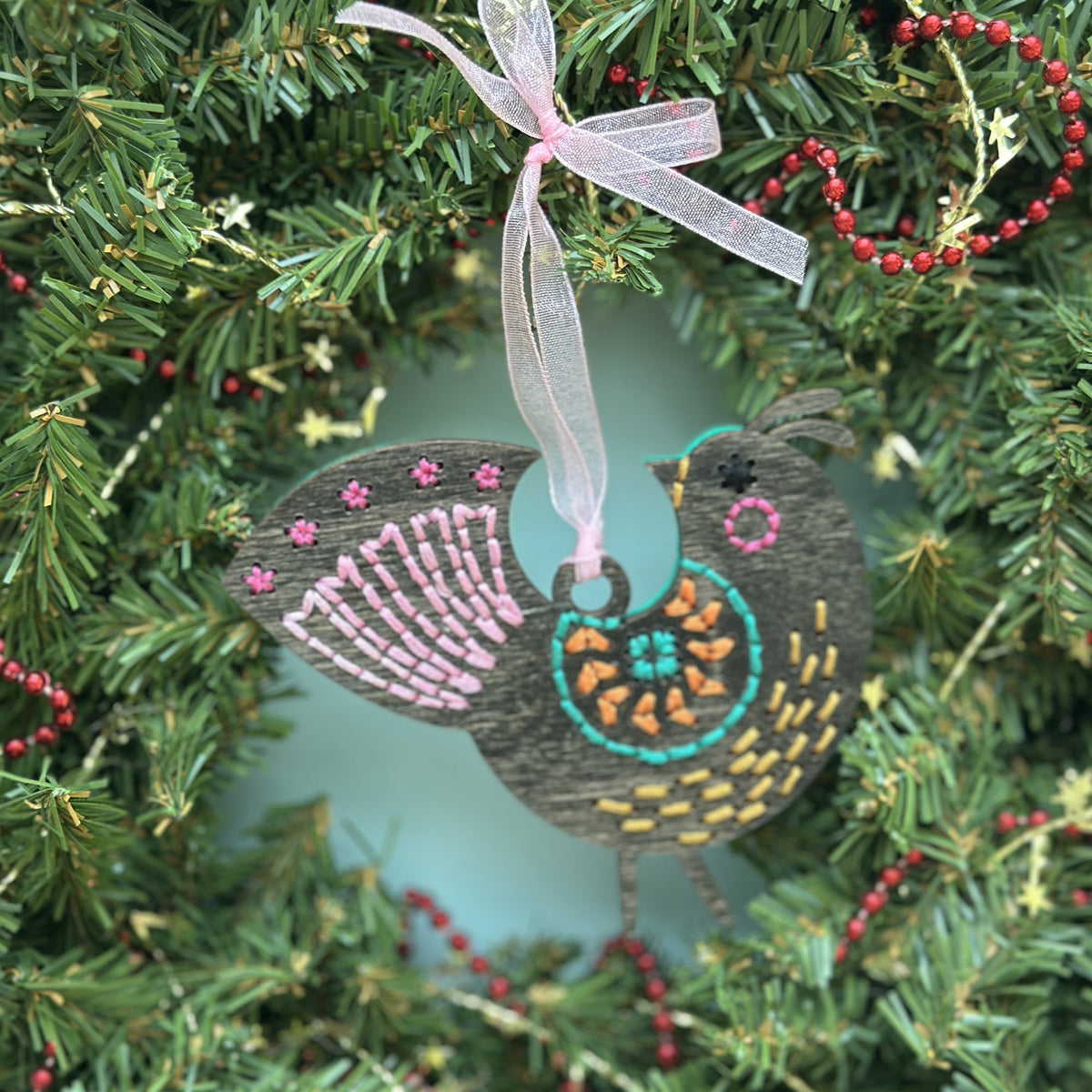 Dark stained medallion folk bird embroidery ornament with a pink ribbon hanging in an evergreen wreath