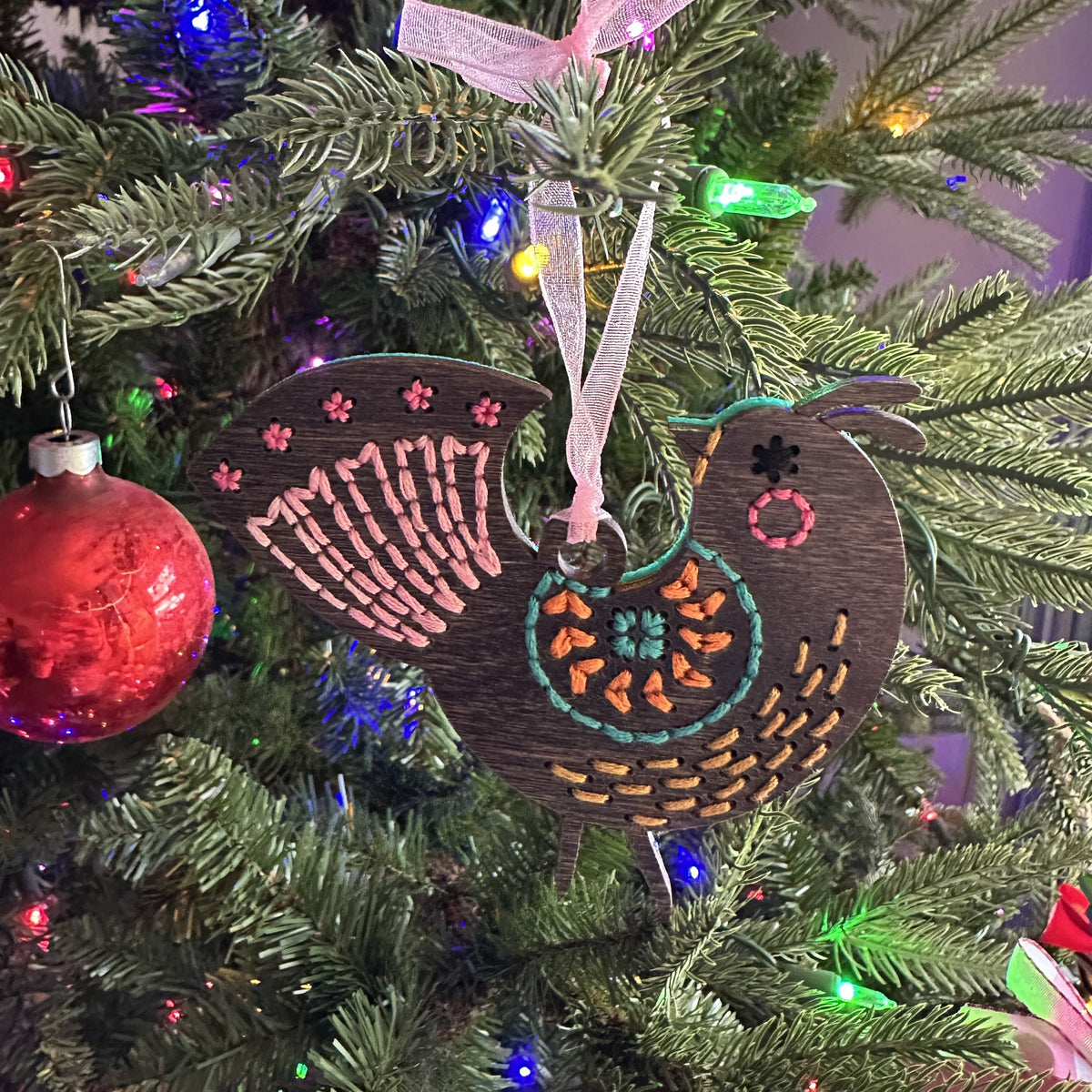 Dark stained medallion folk bird embroidery ornament on a lit Christmas tree with a red ornament