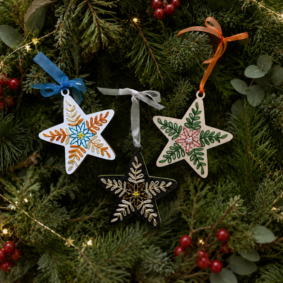 Three folk art star embroidery ornaments in different wood finishes hanging on evergreen branches