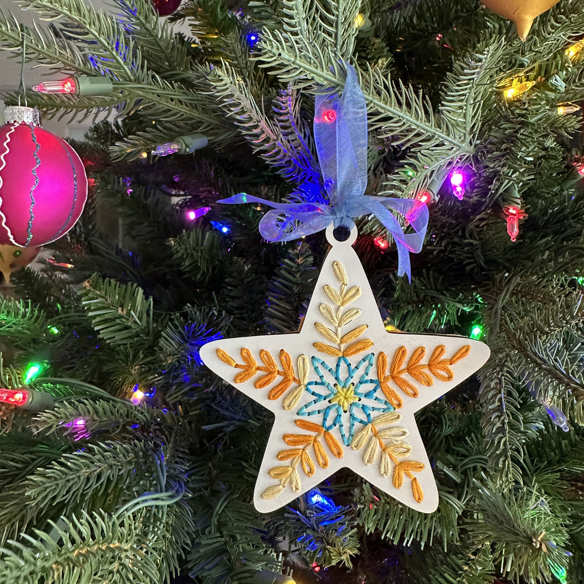 Natural wood folk art star embroidery ornament hanging on a lit Christmas tree