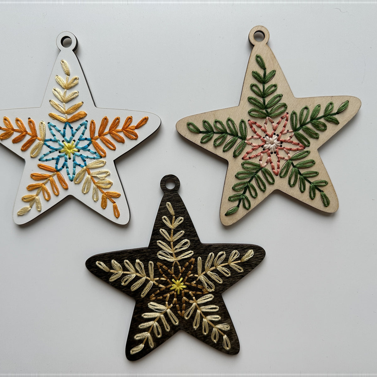 Three folk art star embroidery ornaments in white, natural and dark wood finishes laid flat on a white background