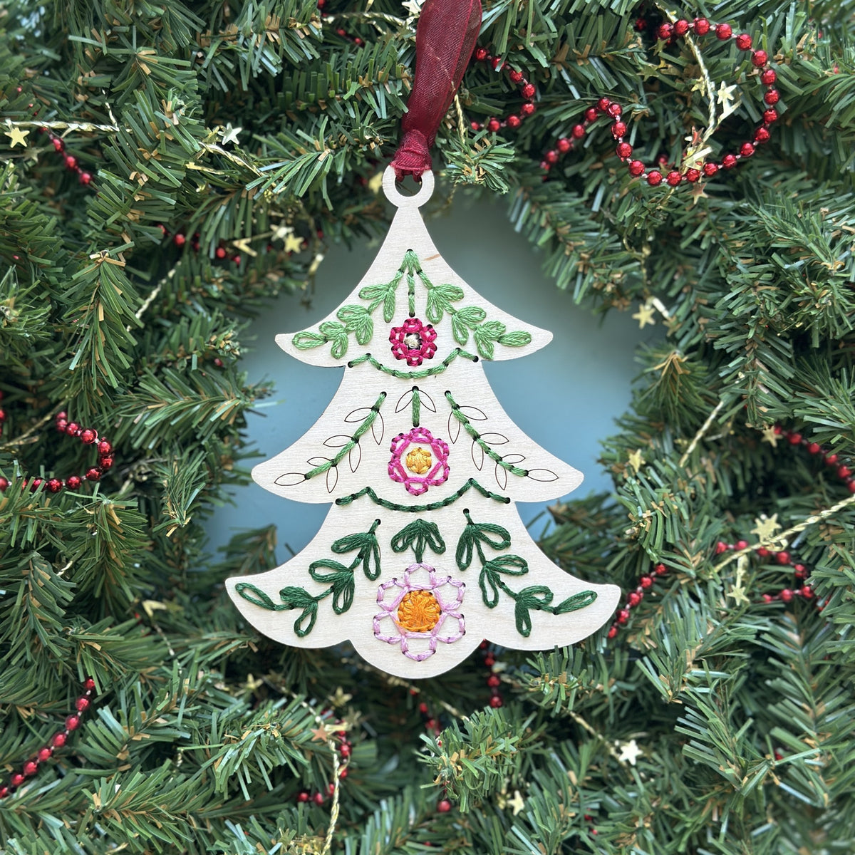 Light wood tree embroidery ornament with green and pink stitching hanging in an evergreen Christmas wreath