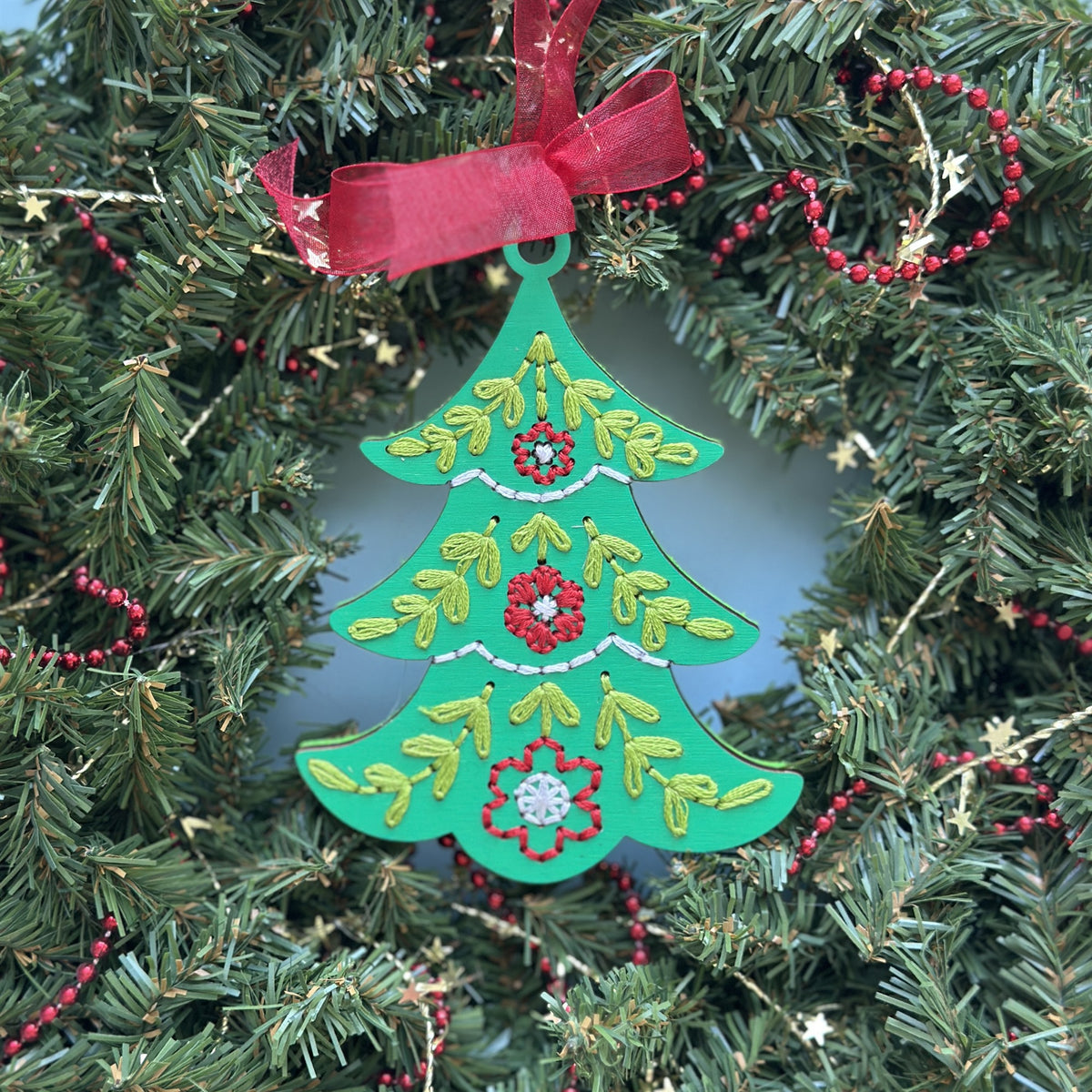 Green tree embroidery ornament with a red ribbon hanging in an evergreen wreath