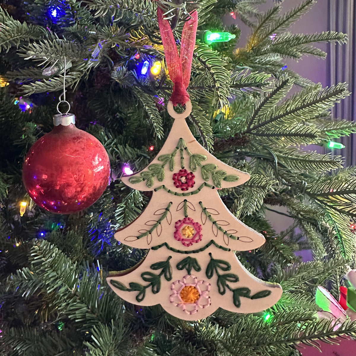 Light wood tree embroidery ornament hanging on a lit Christmas tree with a red ornament