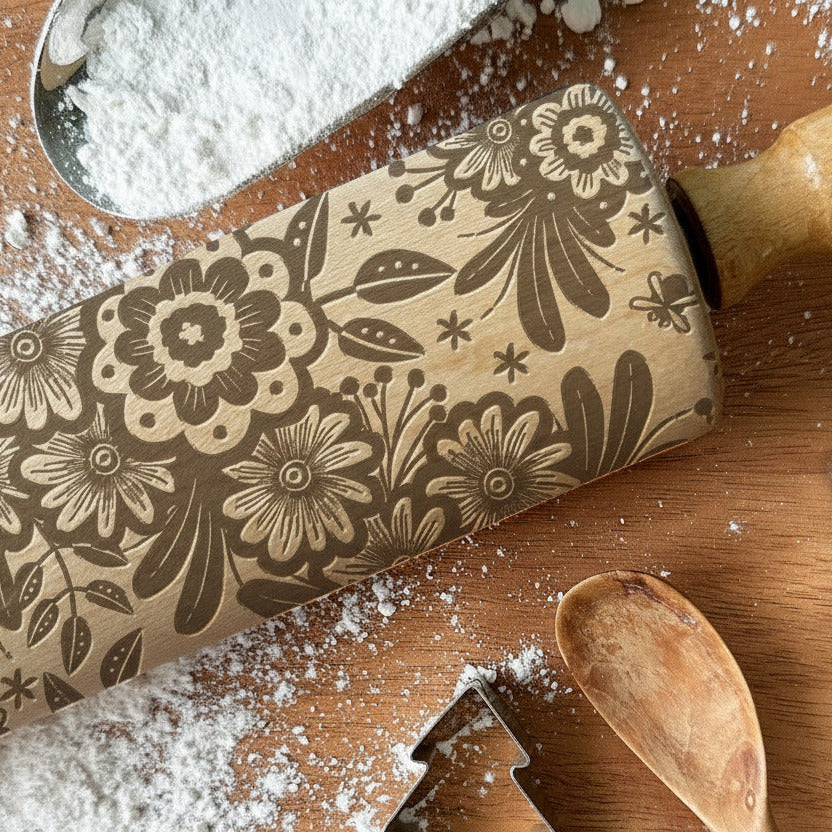 Decorative rolling pin with floral patterns and cookie cutters on a wooden surface with flour.