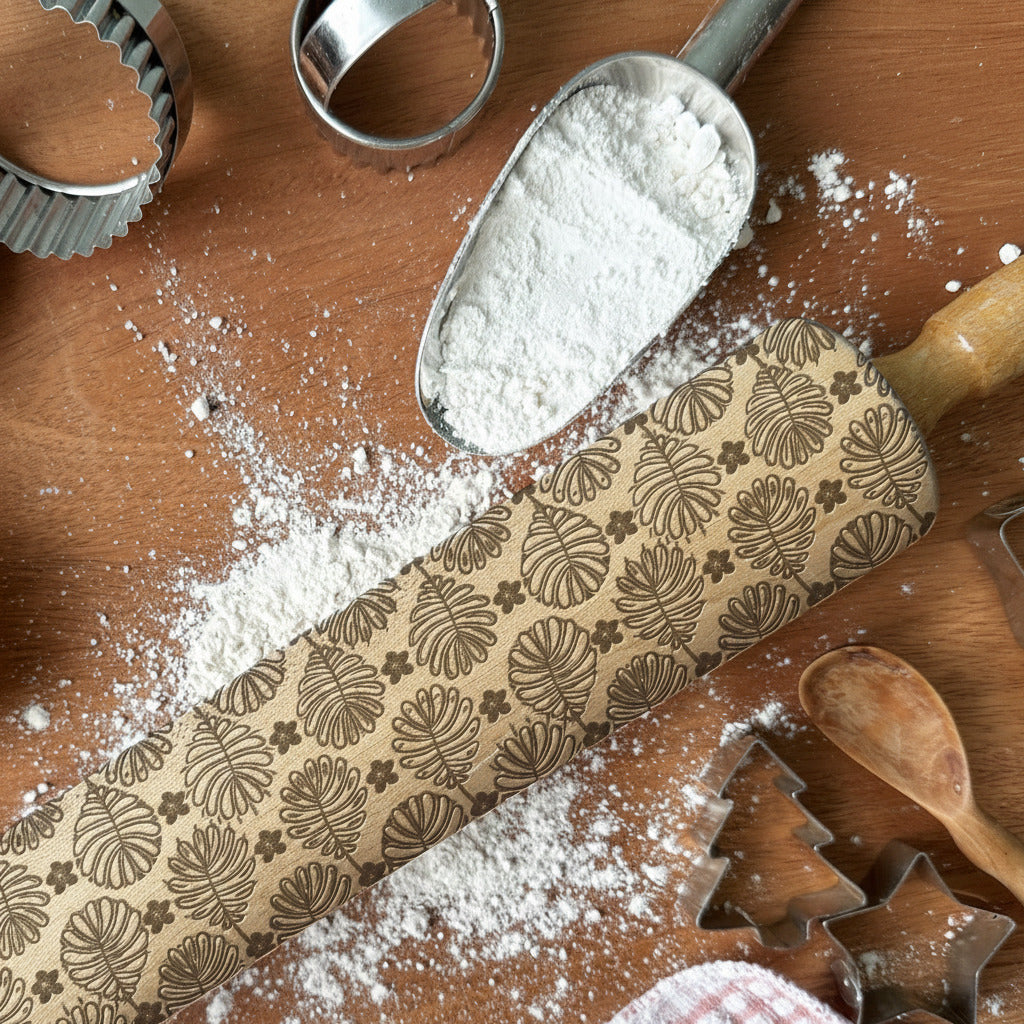 Decorative rolling pin with leaf pattern on a wooden surface with flour and cookie cutters.
