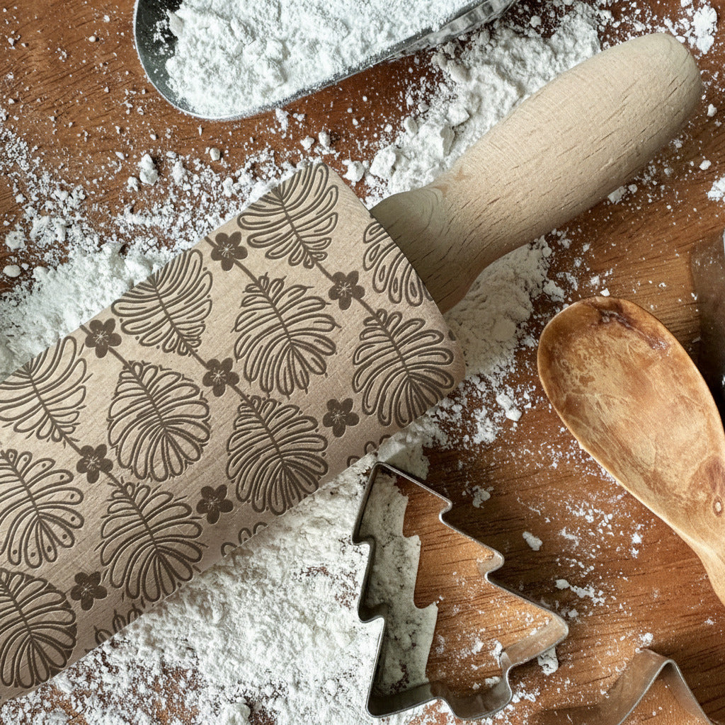 Rolling pin with leaf patterns on a wooden surface with flour, surrounded by cookie cutters.