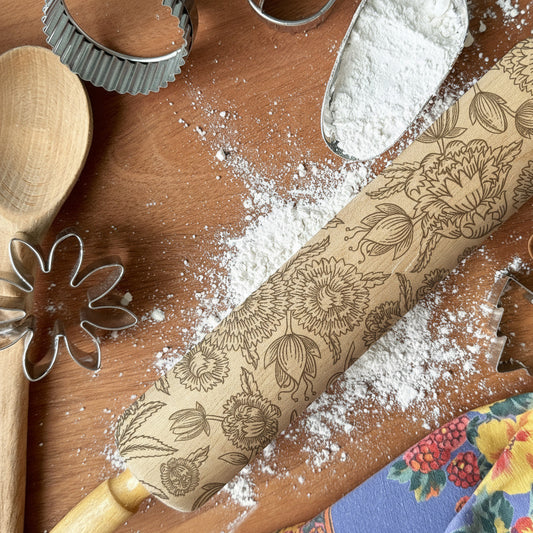Baking tools including a wooden spoon, rolling pin with floral patterns, and cookie cutters on a wooden surface with flour.