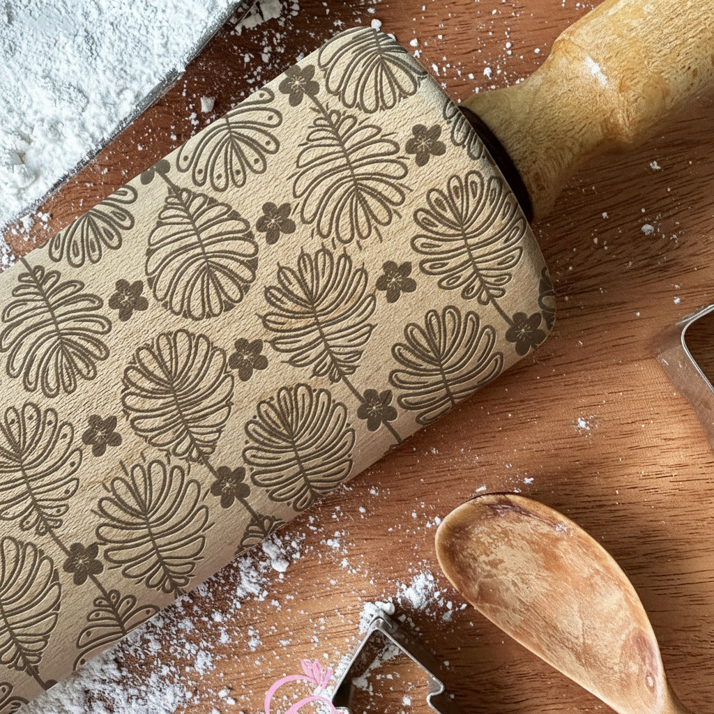 Wooden rolling pin with leaf pattern on a wooden surface with flour, cookie cutter, and branding text.