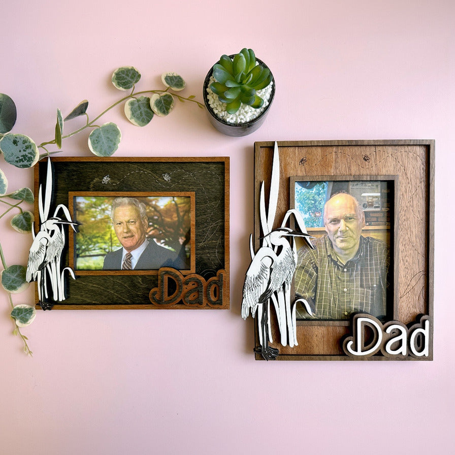 Two wooden picture frames with 'Dad' and heron designs on a pink wall with plants.